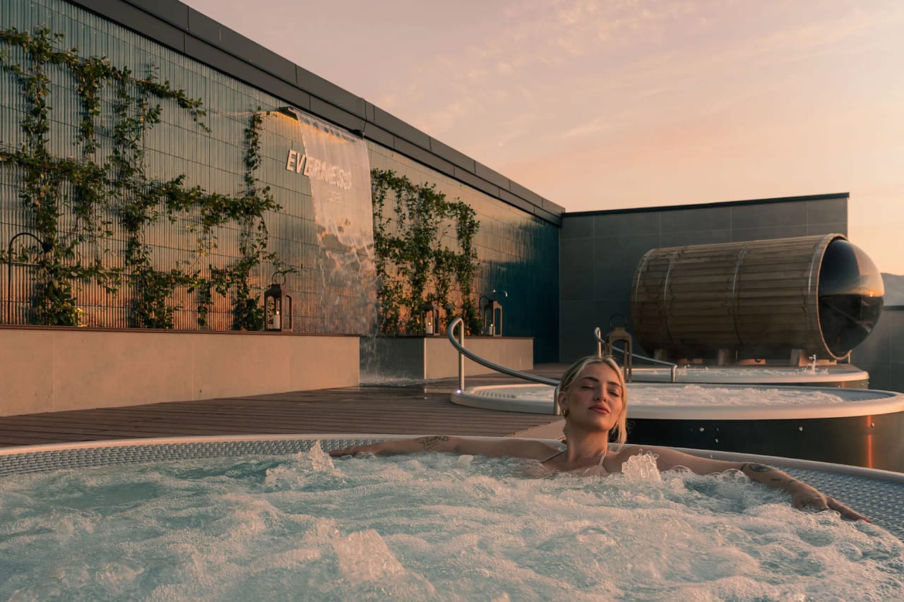 Femme détendue dans le jacuzzi rooftop Everness au coucher de soleil, face au sauna tonneau et au mur végétal